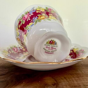 May include: A white Royal Albert Bone China teacup and saucer set. The cup and saucer feature a floral design with pink, yellow, and blue flowers on a burgundy background. The set is trimmed with gold and the text "Garland Series Elegance" is visible.