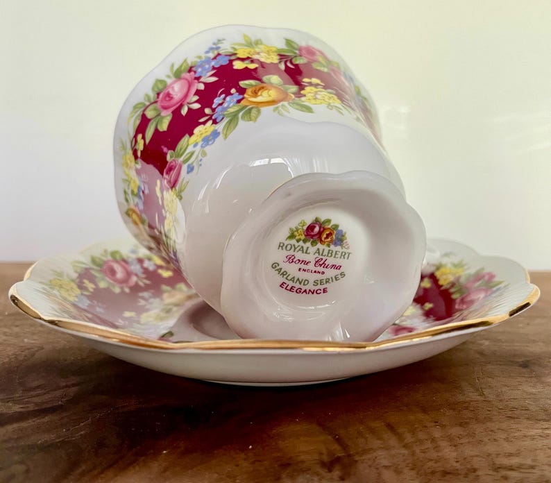 May include: A white Royal Albert Bone China teacup and saucer set. The cup and saucer feature a floral design with pink, yellow, and blue flowers on a burgundy background. The set is trimmed with gold and the text "Garland Series Elegance" is visible.