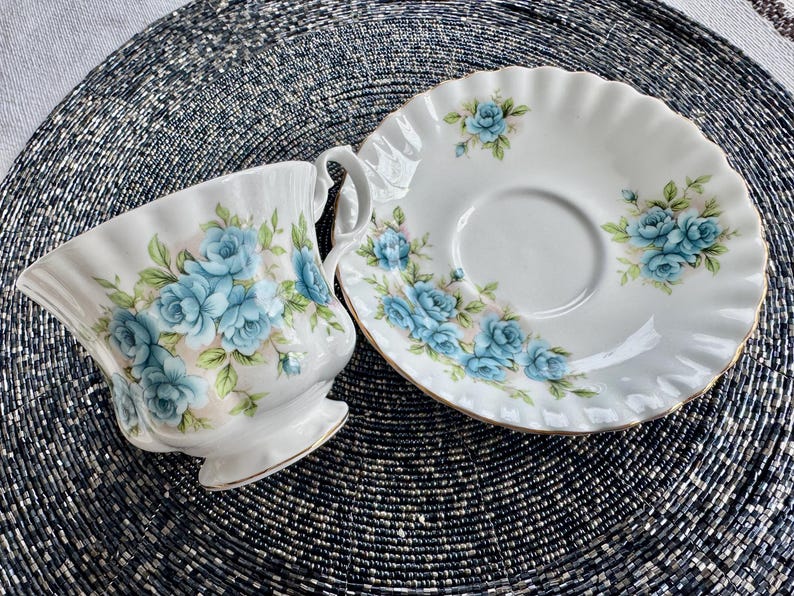 May include: A white teacup and saucer with a blue floral pattern. The cup and saucer have a scalloped edge and a gold trim.
