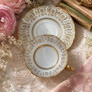 May include: A white porcelain teacup and matching saucer featuring a gold-toned decorative pattern of palmettes and scrolling motifs. The teacup has a scalloped rim and a gold-painted handle, while the saucer displays a wide, ornate gold border.