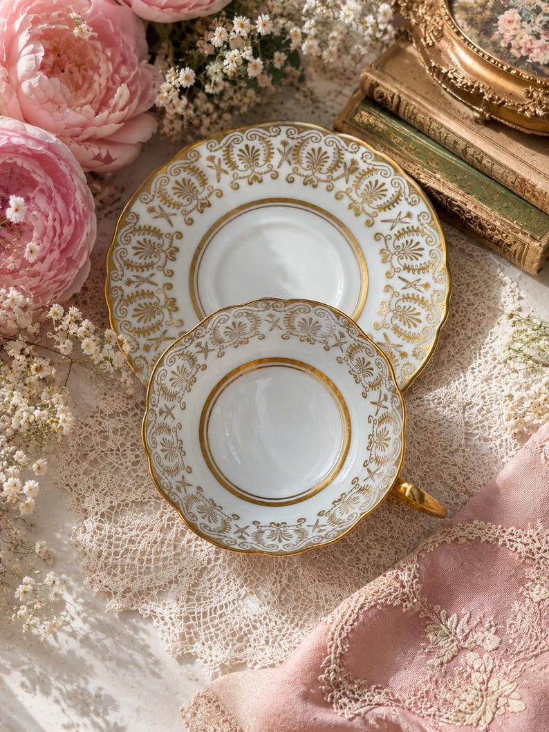 May include: A white porcelain teacup and matching saucer featuring a gold-toned decorative pattern of palmettes and scrolling motifs. The teacup has a scalloped rim and a gold-painted handle, while the saucer displays a wide, ornate gold border.