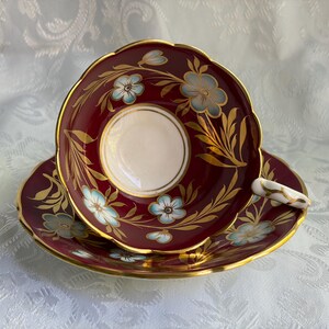 May include: A burgundy teacup and saucer set featuring a white center, scalloped edges, and gold trim. The design includes light blue flowers with gold leaves painted on the deep red surface. The handle is white with gold accents.