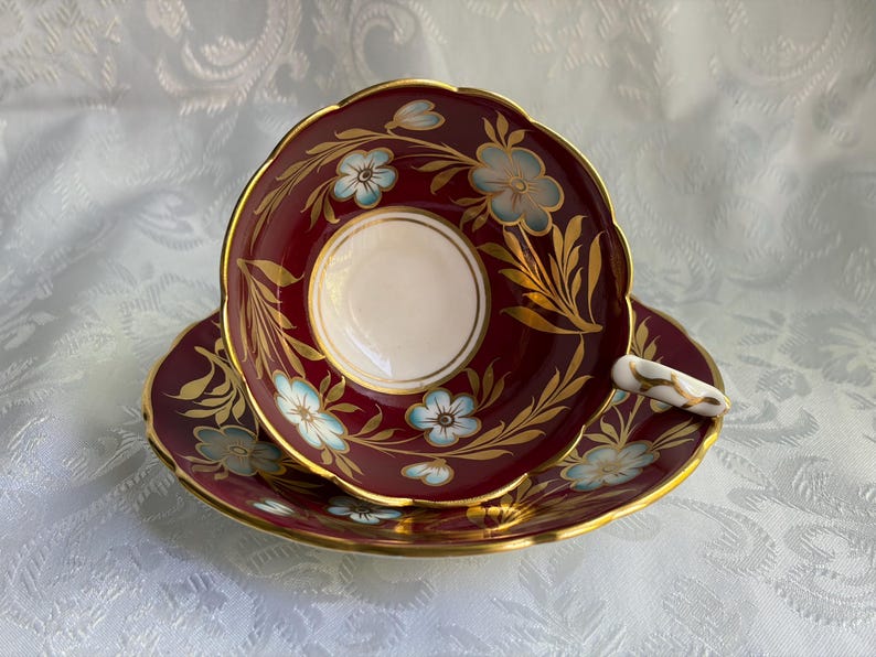 May include: A burgundy teacup and saucer set featuring a white center, scalloped edges, and gold trim. The design includes light blue flowers with gold leaves painted on the deep red surface. The handle is white with gold accents.