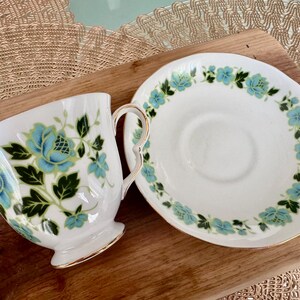 May include: A white teacup and saucer with a floral design. The teacup and saucer have a blue and green floral pattern with gold trim.