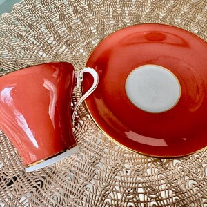 May include: A coral-colored teacup and saucer set with a gold rim. The saucer has a white center circle with a gold rim.