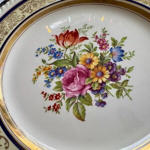 May include: A white porcelain plate with a floral bouquet design. The bouquet features a red tulip, pink rose, and various other colorful flowers. The plate has a gold rim and a decorative border with a navy blue accent.