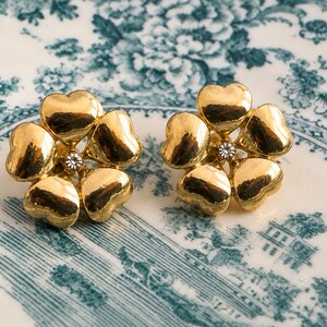 May include: A pair of gold-toned flower-shaped stud earrings. Each earring features five heart-shaped petals surrounding a small, clear crystal. The earrings are displayed on a white and blue floral patterned surface.