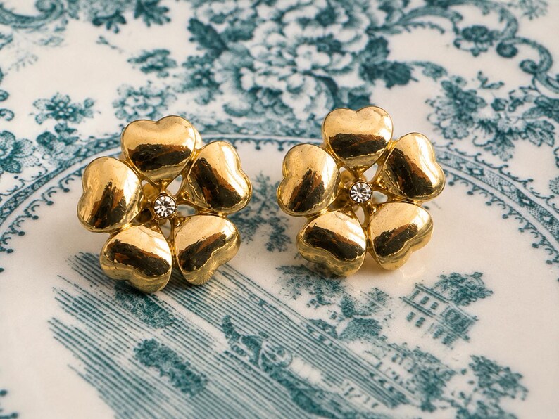 May include: A pair of gold-toned flower-shaped stud earrings. Each earring features five heart-shaped petals surrounding a small, clear crystal. The earrings are displayed on a white and blue floral patterned surface.