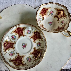 May include: A vintage teacup and saucer set. The white porcelain cup and saucer feature a burgundy and gold ornate design with floral accents. The set is placed on a heart-shaped tray with gold handles.