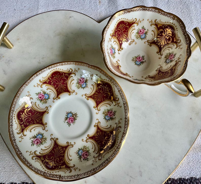 May include: A vintage teacup and saucer set. The white porcelain cup and saucer feature a burgundy and gold ornate design with floral accents. The set is placed on a heart-shaped tray with gold handles.