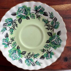 May include: A white teacup saucer with a green and white floral design. The saucer has a scalloped edge and a gold trim.