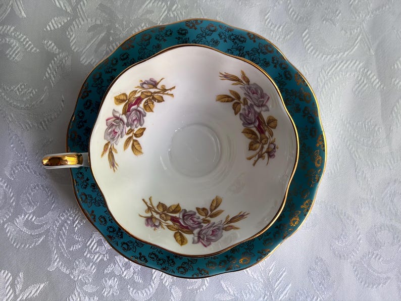 Może przedstawiać: Biały porcelanowy zestaw filiżanki ze spodkiem o falistym brzegu. Spodek jest w kolorze morskim z ciemnym kwiatowym wzorem i złotym wykończeniem. Wnętrze filiżanki zdobią trzy bukiety jasnofioletowych r&oacute;ż z brązowymi liśćmi i złotym rantem.