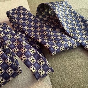 May include: Two neckties featuring a repeating pattern of black and white soccer balls set against a grid of alternating dark blue and light blue squares. The ties are folded and layered, displaying the consistent graphic print across the fabric.