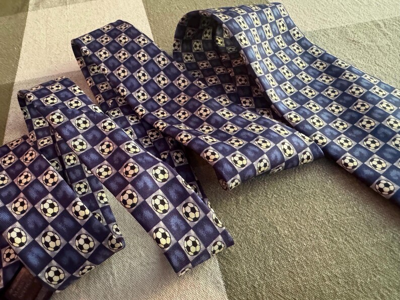 May include: Two neckties featuring a repeating pattern of black and white soccer balls set against a grid of alternating dark blue and light blue squares. The ties are folded and layered, displaying the consistent graphic print across the fabric.