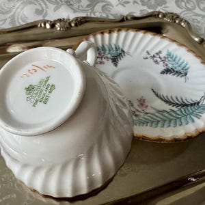 Puede incluir: Juego de taza y platillo de t&eacute; blanco con dise&ntilde;o acanalado. El platillo presenta un patr&oacute;n de helechos y flores con ribete dorado. La taza est&aacute; marcada "Stanley Fine Bone China England EST 1975".