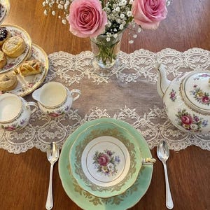 Peut inclure: Un service &agrave; th&eacute; comprenant une tasse et une soucoupe vert menthe avec des filigranes dor&eacute;s, une th&eacute;i&egrave;re blanche, un pot &agrave; lait et un sucrier &agrave; motifs floraux. Un vase en verre contient des roses roses et du gypsophile, avec des p&acirc;tisseries.