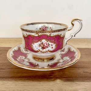 May include: A vintage teacup and saucer set. The teacup and saucer are white with a pink and gold floral design. The teacup has a gold handle and trim. The saucer has a matching design. The set is on a wooden surface.