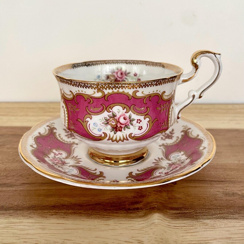 May include: A vintage teacup and saucer set. The teacup and saucer are white with a pink and gold floral design. The teacup has a gold handle and trim. The saucer has a matching design. The set is on a wooden surface.