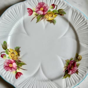 May include: White, scalloped-edge plate with a light blue rim. The plate features a floral design with pink, yellow, and red flowers and green leaves. The plate has a ridged texture around the edge.