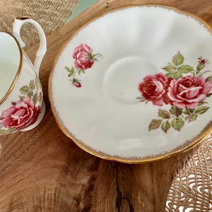 Pu&ograve; includere: Una tazzina da t&egrave; e un piattino bianchi con un bordo dorato e un motivo floreale con rose rosa. Il piattino ha un bordo ondulato.