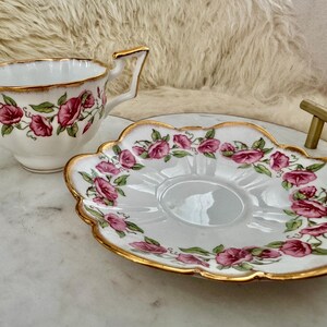 Puede incluir: Juego de taza y platillo blancos con un estampado floral de rosas rosas y hojas verdes. Los bordes est&aacute;n adornados con dorado. La taza tiene un asa dorada y el platillo tiene un borde festoneado.