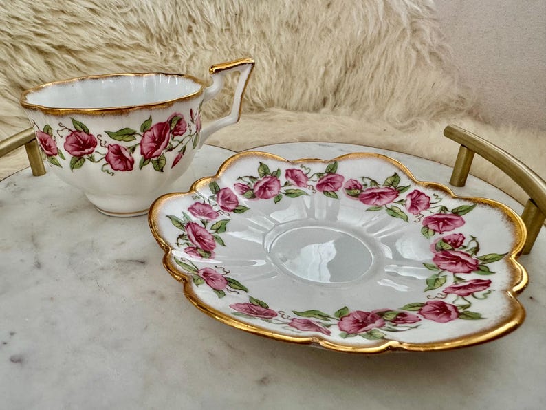 Puede incluir: Juego de taza y platillo blancos con un estampado floral de rosas rosas y hojas verdes. Los bordes est&aacute;n adornados con dorado. La taza tiene un asa dorada y el platillo tiene un borde festoneado.