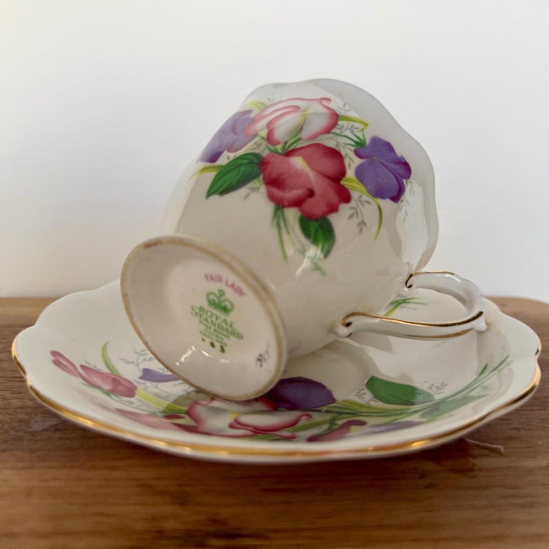 Może przedstawiać: Zestaw filiżanki i spodka do herbaty w stylu vintage. Biała porcelanowa filiżanka i spodek mają kwiatowy wz&oacute;r z czerwonymi, fioletowymi i zielonymi akcentami. Zestaw ma złote wykończenia, a filiżanka jest oznaczona "Royal Standard Fair Lady".