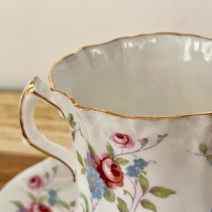 May include: Close-up of a white teacup and saucer with a floral pattern of red, pink, and blue flowers. The rim and handle are trimmed with gold. The teacup is on a wooden surface.