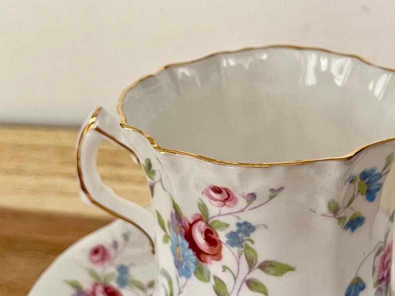 May include: Close-up of a white teacup and saucer with a floral pattern of red, pink, and blue flowers. The rim and handle are trimmed with gold. The teacup is on a wooden surface.