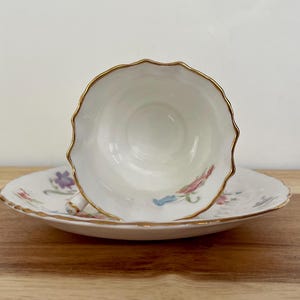 May include: White teacup and saucer set with a scalloped gold rim. The teacup is positioned above the saucer, revealing a floral pattern in pink, purple, and blue. The set is on a wooden surface.