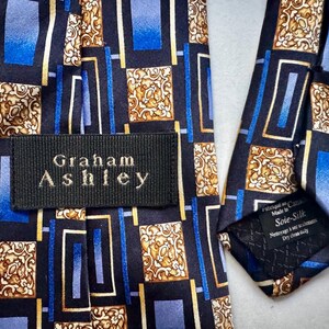 May include: A necktie featuring a black background with a geometric pattern of blue rectangles and gold ornate square motifs. A black fabric label with the white text Graham Ashley is stitched onto the front, and a care tag reads Soie-Silk.