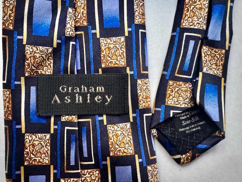 May include: A necktie featuring a black background with a geometric pattern of blue rectangles and gold ornate square motifs. A black fabric label with the white text Graham Ashley is stitched onto the front, and a care tag reads Soie-Silk.
