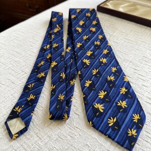 May include: A blue tie with a repeating pattern of yellow floral designs and diagonal stripes. The tie is made of a smooth fabric and is displayed on a white surface. A brown box is visible in the background.