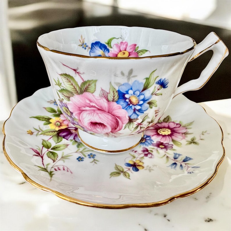 May include: A white teacup and saucer set with a floral pattern. The cup and saucer are decorated with pink, blue, yellow, and purple flowers. The edges of the cup and saucer are trimmed with gold.