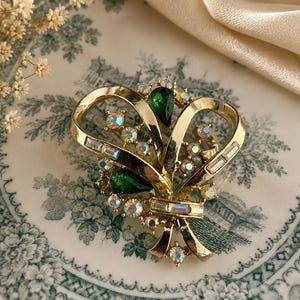 May include: A vintage gold-tone brooch with a bow design. The brooch features green teardrop-shaped stones and iridescent rhinestones. The brooch is set against a patterned plate and a cream-colored fabric.