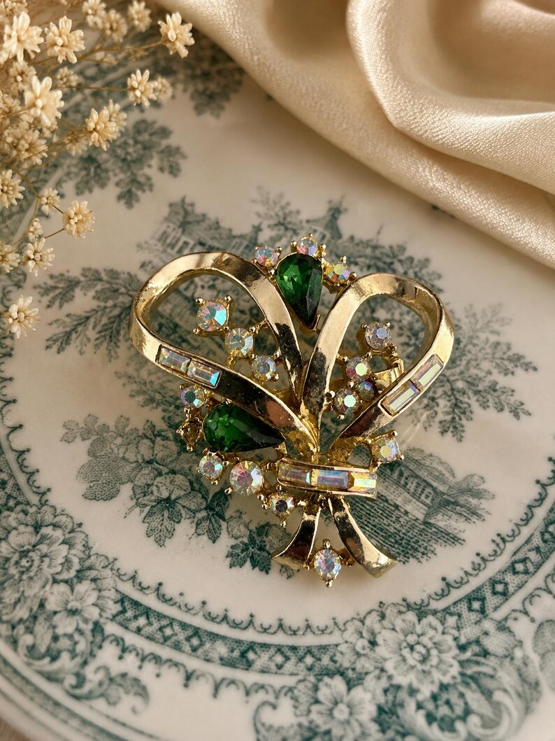 May include: A vintage gold-tone brooch with a bow design. The brooch features green teardrop-shaped stones and iridescent rhinestones. The brooch is set against a patterned plate and a cream-colored fabric.