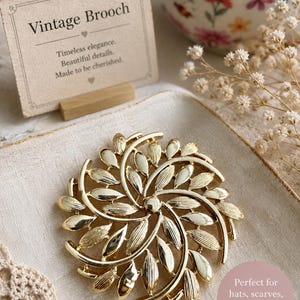 May include: A gold-toned vintage brooch with a leaf design. The brooch is circular with a central point and radiating leaves. The card next to it reads "Vintage Brooch" and "Timeless elegance. Beautiful details. Made to be cherished."