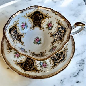 May include: An ornate teacup and saucer set. The white porcelain features black and gold filigree designs, with accents of pink and blue floral patterns. The rim and handle are trimmed in gold, creating an elegant aesthetic.