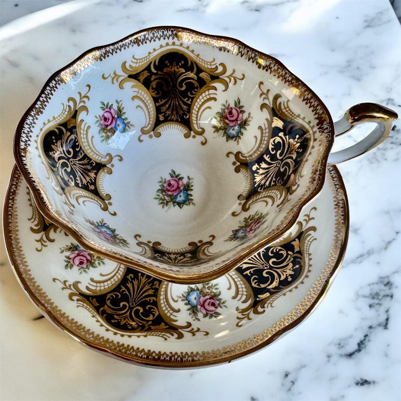 May include: An ornate teacup and saucer set. The white porcelain features black and gold filigree designs, with accents of pink and blue floral patterns. The rim and handle are trimmed in gold, creating an elegant aesthetic.