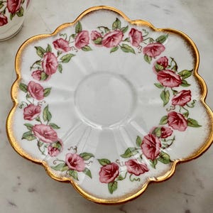 Puede incluir: Platillo blanco con borde festoneado y ribete dorado. El platillo est&aacute; decorado con un c&iacute;rculo de flores rosas y rojas con hojas verdes. Se ve parcialmente una taza a juego.