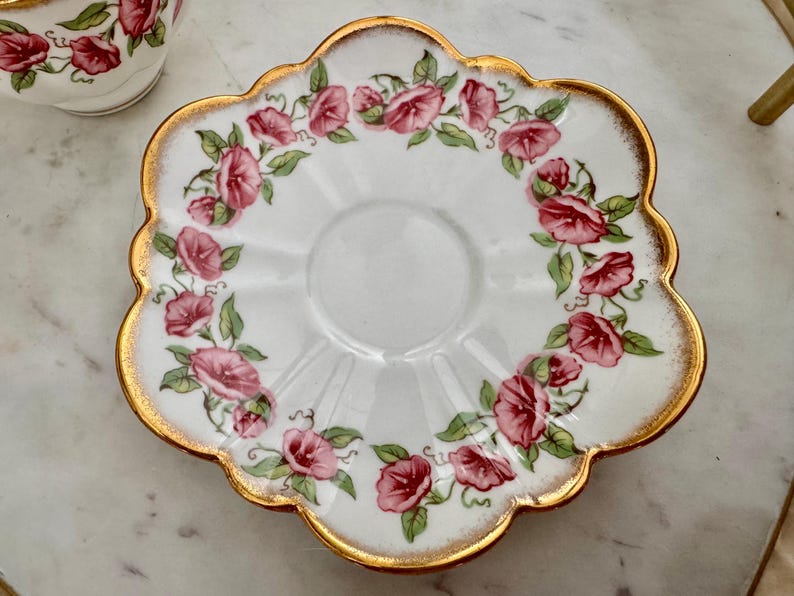 Puede incluir: Platillo blanco con borde festoneado y ribete dorado. El platillo est&aacute; decorado con un c&iacute;rculo de flores rosas y rojas con hojas verdes. Se ve parcialmente una taza a juego.