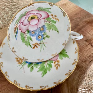 May include: A white teacup and saucer set with a floral design. The cup and saucer feature a pink flower with blue and yellow accents, surrounded by green leaves and gold trim.