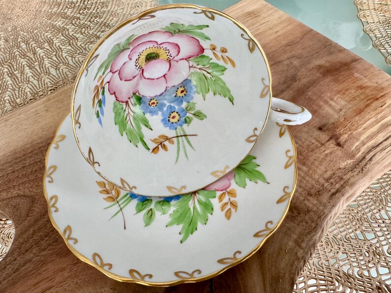 May include: A white teacup and saucer set with a floral design. The cup and saucer feature a pink flower with blue and yellow accents, surrounded by green leaves and gold trim.