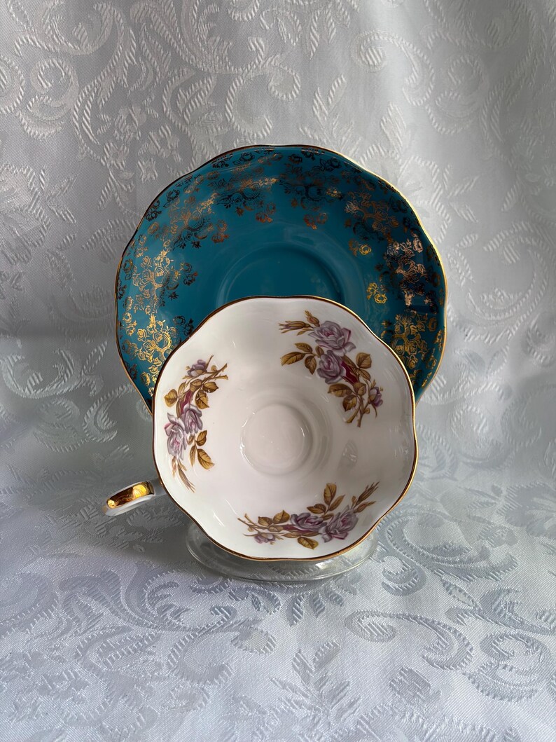 Może przedstawiać: Biała porcelanowa filiżanka do herbaty ze spodkiem o falistych brzegach. Filiżanka ozdobiona jest motywami fioletowych r&oacute;ż, natomiast spodek posiada głębokie turkusowe tło z misternym złotym wzorem filigranowym na całej powierzchni.
