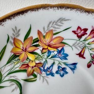 May include: Close-up of a white saucer with a gold rim. The saucer features a floral design with yellow, orange, red, and blue flowers.