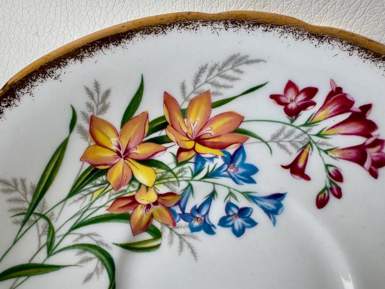 May include: Close-up of a white saucer with a gold rim. The saucer features a floral design with yellow, orange, red, and blue flowers.