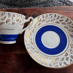 May include: A white teacup and saucer set with a blue and gold design. The teacup has a blue band around the top and gold leaf designs. The saucer has a blue center circle with a gold rim and gold leaf designs around the edge.