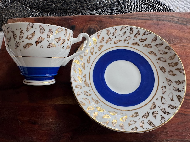 May include: A white teacup and saucer set with a blue and gold design. The teacup has a blue band around the top and gold leaf designs. The saucer has a blue center circle with a gold rim and gold leaf designs around the edge.