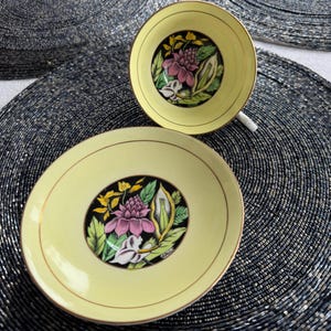 May include: A yellow teacup and saucer with a black and gold floral design. The flowers are pink, white, and yellow.