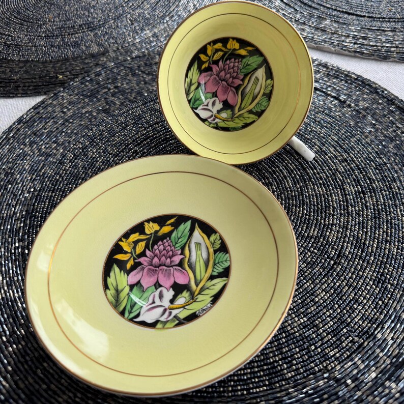 May include: A yellow teacup and saucer with a black and gold floral design. The flowers are pink, white, and yellow.